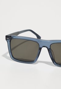 Óculos de sol transparentes azuis com lentes cinza escuro, design de armação quadrada e braçadeiras finas. Destacam-se pelos acessórios minimalistas na armação.