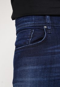 Vaqueros de mezclilla azul oscuro con un ajuste slim, un ligero desgaste, acentos en los bolsillos frontales con botones negros y un diseño clásico de cinco bolsillos.