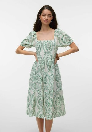 Kvinde med langt mørkt hår iført en hvid midi-kjole med grøn blomsterbroderi og puffede ærmer, stående med hænderne på hofterne.