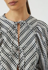 Blusa a scacchi bianca e nera con chiusura a fiocco, caratterizzata da maniche a palloncino e scollatura a chiave, realizzata in tessuto leggero e tessuto.