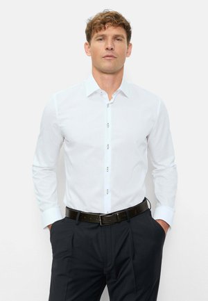 Mann mit lockigem Haar trägt ein weißes Hemd mit langen Ärmeln, das in dunkle Hosen mit schwarzem Gürtel gesteckt ist, steht mit den Händen in den Taschen.