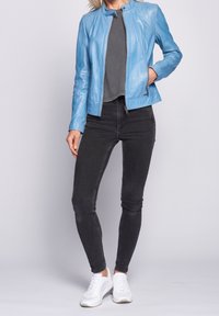 Kvinde iført en lyseblå læderjakke, mørkegrå t-shirt, sorte skinny jeans og hvide sneakers, stående foran en ensfarvet baggrund.