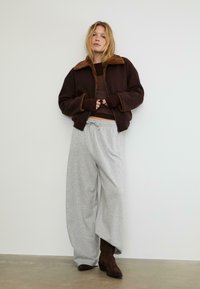 Giacca bomber marrone con colletto in pelliccia beige, indossata sopra un top fitted marrone, abbinata a pantaloni larghi grigi chiari e stivali marroni.