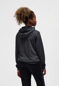 Junge Person mit lockigem, hochgebundenem Haar, trägt eine dunkelgraue Kapuzenjacke und schwarze Hosen mit gemusterten Seitenstreifen, von hinten gesehen.