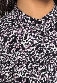 Nahaufnahme einer Bluse mit hohem Kragen, schwarzen Knöpfen und einem abstrakten Muster in Schwarz, Weiß und Lila. Geflochtenes Haar ist seitlich sichtbar.