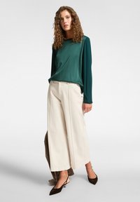 Pitkähihaista vihreää paitaa on paritettu kerma- värisillä leveälahkeisilla housuilla. Mallilla on päällään tummanruskeat kärjelliset kengät, jotka korostavat sileää kangasta ja rentoa istuvuutta.