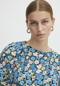 Blusa floreale con sfondo nero, caratterizzata da fiori blu e rosa. Collo rotondo, tessuto morbido e maniche a palloncino corte. Orecchini ad anello dorati.