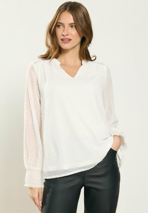 Frau mit hellbraunen Haaren, die eine weiße, strukturierte Bluse mit langen Ärmeln und schwarze Hose trägt, steht mit einer Hand in der Hosentasche und schaut nach rechts.