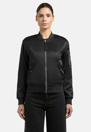 Vrouw draagt een zwarte bomberjack met ritssluiting, zakken en lange mouwen, gecombineerd met een zwarte broek, staand tegen een effen achtergrond.