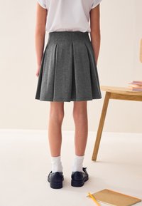 Jupe plissée grise avec une ceinture haute, fabriquée dans un tissu doux. Portée avec des chaussures noires et des chaussettes blanches. Vue de dos.