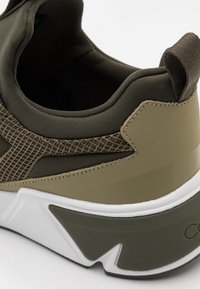 Närbild av en kuddad klack på en sneaker i olivgrönt och beige, med vadderad krage, tygdragflik, texturerad sidopanel och tjock vit och olivfärgad sula.