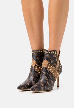 Stivaletti alla caviglia con tacchi alti, neri e marroni, con punte appuntite, borchie dorate e motivo monogramma ripetuto, indossati su gambe scoperte.