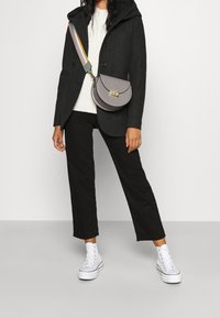 Bolso bandolera gris con forma curva, con herrajes dorados y una correa a rayas. Llevado sobre un suéter blanco y pantalones negros, con zapatillas blancas.