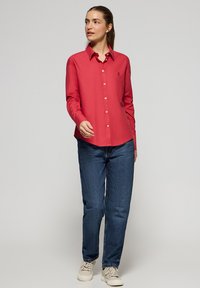Camicia rossa a maniche lunghe con colletto, abbinata a jeans blu di denim e scarpe beige, presentata su uno sfondo neutro.