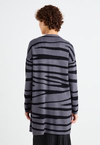 Grå cardigan med svart zebra-stripe mønster, lange ermer og avslappet passform. Myk tekstur med en middels lang design.