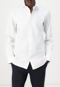 Man in een witte button-up shirt met handen in de zakken en marineblauwe gestreepte broek tegen een eenvoudige witte achtergrond.