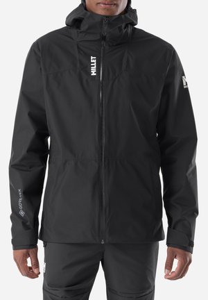 Man wearing a black Gore-Tex waterproof jacket with a hood, zipped up, with white brand logos on chest, sleeve, and pants.