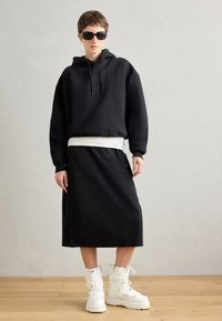 Svart oversized hoodie med snoddar fram, över en vit topp, tillsammans med en svart midikjol och kraftiga vita stövlar.