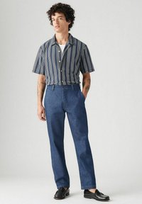 Lyhythihainen raidallinen paita, jossa on napitus, yhdistetty suoralahkeisiin farkkuihin; malli käyttää mustia loafereita ja seisoo neutraalin taustan edessä.