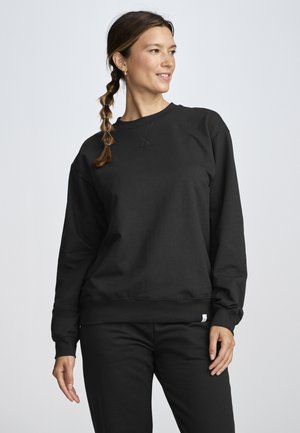 Frau mit geflochtenem Haar, trägt einen schlichten schwarzen Sweatshirt und schwarze Hose, steht vor einem hellgrauen Hintergrund und schaut nach rechts.