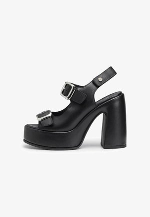 Sandales à talons hauts noires avec une plateforme épaisse, dotées de deux brides à boucle en argent, bout ouvert et design slingback. Texture en cuir lisse.