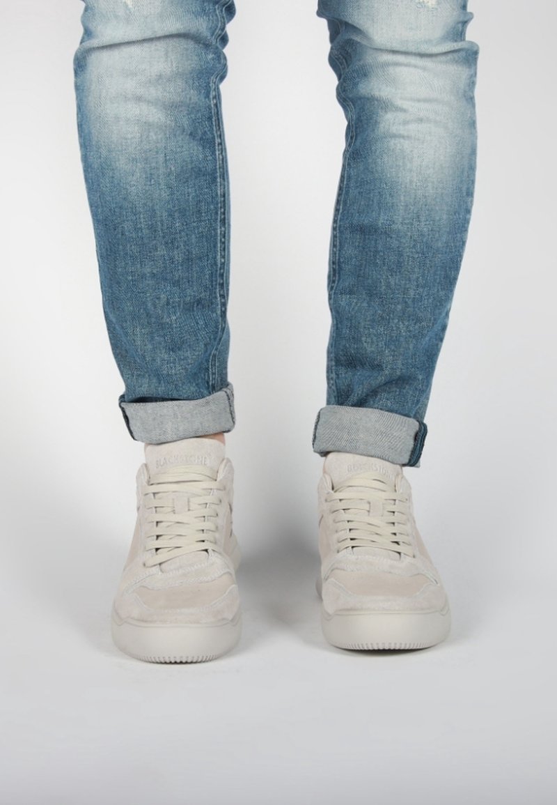 Personne portant un jean bleu délavé retroussé et des baskets blanches à lacets, debout sur un fond clair uni.