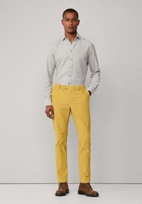 Harmaa napillinen paita pitkillä hihoilla, yhdistettynä keltaisiin chinopö pantsseihin ja ruskeisiin nahkabootseihin. Silkkiset kankaat ja säädetty istuvuus.