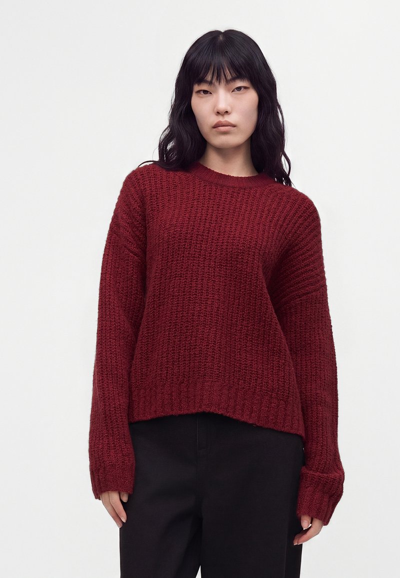 Jeune femme aux longs cheveux noirs portant un pull texturé rouge foncé et un pantalon noir sur un fond clair uni.
