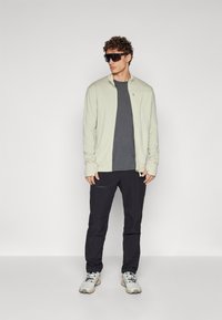 Light beige zip-up jacket over a dark gray t-shirt, paired with black pants and light gray sneakers. Model wears oversized black sunglasses.