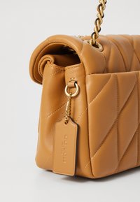 Kviltad beige läderhandväska med rullad topp, kedjestropp och guldfärgad hårdvara. Innehåller en tagg med präglad branding och diagonala sömmar.