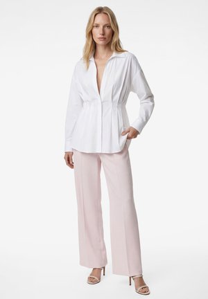 Femme debout portant un chemisier blanc à manches longues avec un décolleté plongeant, un pantalon large rose clair, et des sandales à talons beiges sur fond blanc.