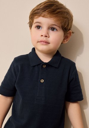 Niño pequeño con cabello castaño claro que lleva una camiseta tipo polo azul marino oscuro, mirando ligeramente hacia arriba y hacia un lado sobre un fondo liso.