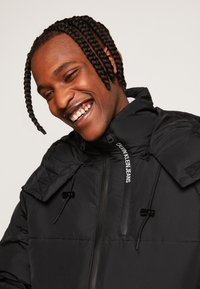 Giubbotto imbottito nero con colletto alto, chiusura con zip e cordini regolabili. Presenta dettagli trapuntati sulle spalle e un logo lungo la zip.