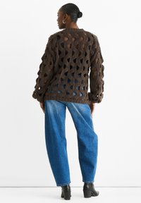 Brown knit sweater with large cut-out pattern, long sleeves, and ribbed cuffs. Paired with loose blue jeans and black heeled boots.