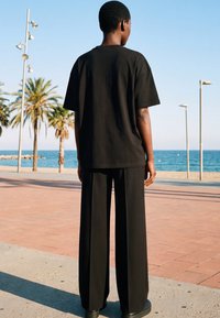 Person in schwarzem Hemd und weitem Hosen steht auf einer gepflasterten Promenade, dem Meer zugewandt, mit Palmen und Straßenlaternen in der Nähe.