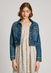 Chaqueta de mezclilla en azul oscuro con un diseño recortado, que cuenta con cierre de botones, bolsillos en el pecho y costuras en contraste, combinada con un vestido floral.