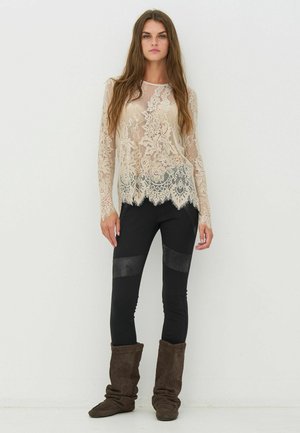 Beiger Spitzenlangarmshirt mit gewelltem Saum über schwarzen Leggings mit lederähnlichen Akzenten. Das Modell trägt braune Wildlederstiefel.