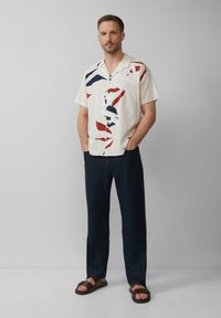 Kurzarmhemd in Creme mit abstrakten roten und blauen Mustern, kombiniert mit dunkler Jeans und braunen Sandalen.