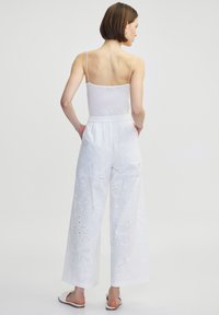 Un pantalon large blanc présente une broderie œillet avec des motifs floraux, une taille élastique et une texture lisse. Porté avec un top blanc à bretelles spaghetti.