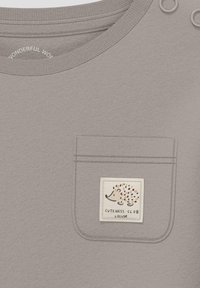 Graues Baumwollhemd mit Rundhalsausschnitt, ausgestattet mit einer kleinen Tasche und einem Igel-Patch, das mit "Cuteness Club" in orangefarbenen Akzenten versehen ist.