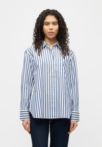 Γυναίκα με σγουρά μαλλιά που φοράει μπλε και άσπρο πουκάμισο με κάθετες ρίγες και σκούρο τζιν, στέκεται μπροστά από ένα απλό λευκό φόντο.