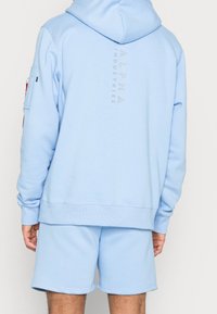 Felpa leggera azzurra con coulisse, caratterizzata da un logo lucido "ALPHA INDUSTRIES" sulla schiena e polsini a costine, abbinata a pantaloni corti coordinati.