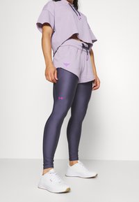 Žena v levandulové zkrácené mikině, odpovídajících šortkách s logem býka, tmavě fialových legínách s růžovým logem a bílých teniskách stojící proti bílé zdi.