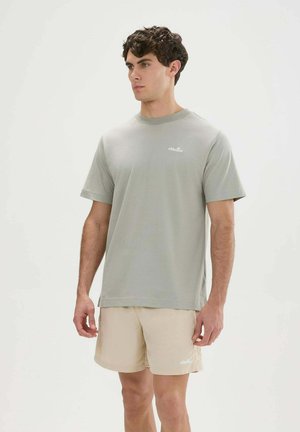 Jonge man met een lichtgrijs shirt met korte mouwen en beige korte broek, staand en lichtjes opzij kijkend tegen een effen achtergrond.