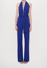 Niebieski kombinezon na szyję z głębokim dekoltem w kształcie litery V i szerokimi nogawkami. Posiada pasek w talii i metaliczny łańcuch jako ozdobę. Gładka faktura tkaniny.