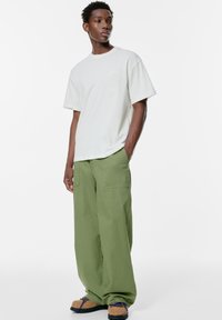 Maglietta di cotone grigia chiara con colletto a girocollo e dettagli con logo, abbinata a pantaloni verdi svasati e larghi con pieghe e scarpe slip-on marroni con lacci colorati.