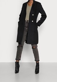 Abrigo de mezcla de lana negro con cuello alto y cierre de doble botón. Combinado con una blusa de canalé oliva, jeans grises desgastados y botas de tobillo negras.