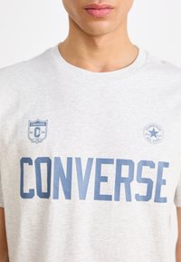 Hele halli puuvillane T-särk ümmarguse kaelusega, millel on sinise "CONVERSE" kiri ja logo aktsendid rinnal.