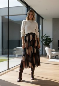 Kremowy dziergany sweter, czarny pasek, plisowana spódnica z dużymi brązowymi i beżowymi wzorami oraz brązowe botki na obcasie. Nowoczesne wnętrze w tle.