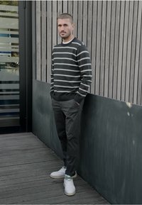 Grijze gestreepte trui met zwart-witte strepen, gecombineerd met donkere broek en witte sneakers. Staand tegen een houten muur buiten.